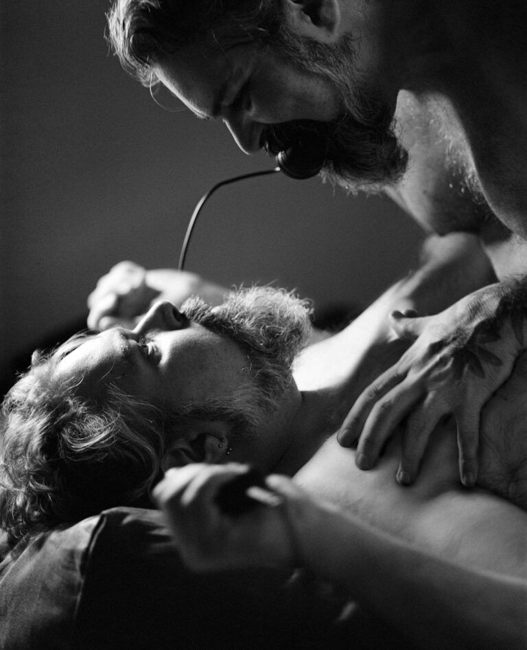 In a photograph, two bodies face each other while in bed: one is pinned down by the other, but the one on top is being gagged by the camera’s remote control, which is also in the one on the bottom’s hand.