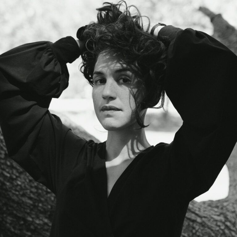 A close up grayscale photo of artist Natalia Lassalle-Morillo standing in front of a tree with her hands behind her head. She looks at the camera with a neutral expression.