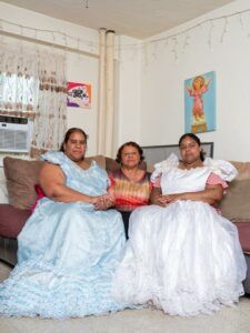 Three women, two with fluffy dresses and one with a patterned blouse sits on a couch at home.