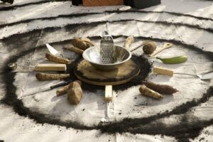 A bowl, grater, and various tools mixed with root vegetables and platanos are set in a radial position in the middle of concentric charcoal circles on canvas.