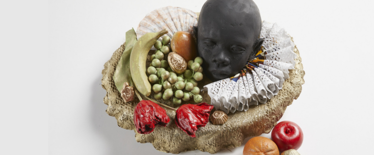 Joey Quinones's artwork Still Life for Black Peter 24" x 18" x 16" Brass, Dutch wax print, lace, paint Photo: Kohler Co: Courtesy of John Michael Kohler Arts Center. a round grouping of flowers fruit lace, shell with a black figures head nestled in the still life.