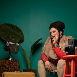 Artist Sadie Woods, photo credit Seed Lynn. Artist Sadie woods sits in front of a green wall, her face resting in her hand. Nearby there is a vintage camera, fan, plant, and yellow suitcase.