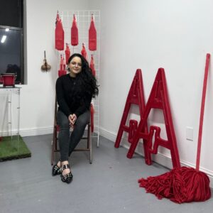 Artist Jordan Corine Cruz sits in a chair dressed in all black in her studio surrounded by red sculptures.