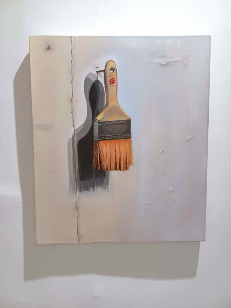 A painting of an orange-stained paintbrush hanging from a nail driven into a white studio wall.